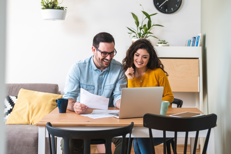 Man and Woman sitting at table and smiling at a computer while going through paperwork.