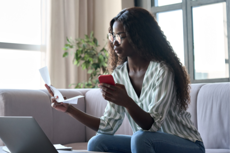 Young woman wearing glasses looking at receipt and holding cell phone.