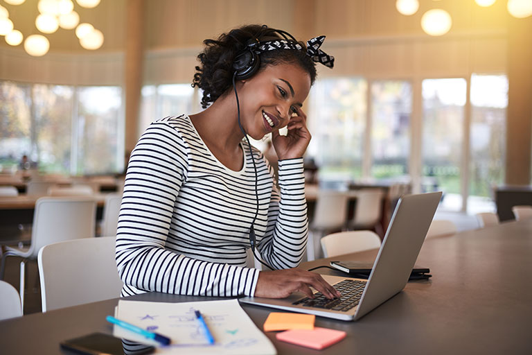 Beautiful young African American woman with short curly hair, listening with headphones and working on her computer.
