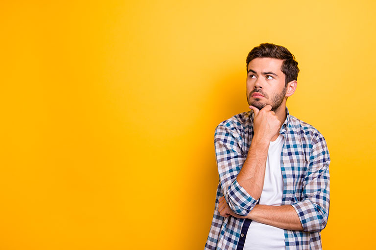 Young man, brown hair and facial hair in plaid shirt, looking to the left with hand on his chin in front of yellow background