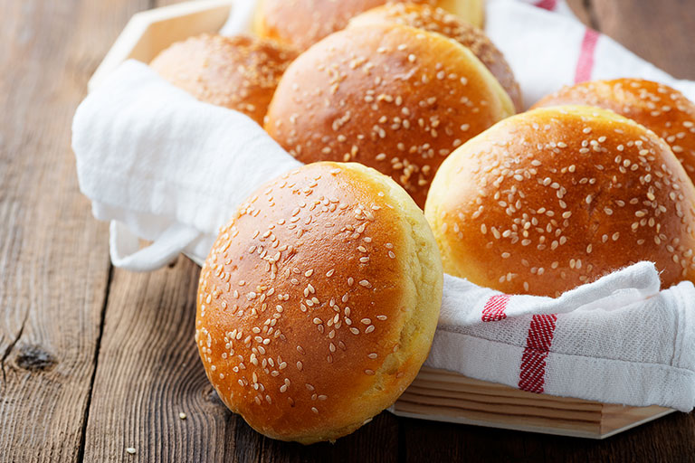 Basket lined with red striped towel filled with hamburger buns with sesame seeds on rustic wooden table