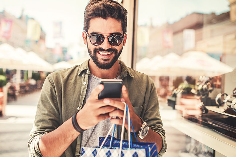 Smiling bearded man wearing sunglasses, walking outside looking at cell phone holding shopping bags