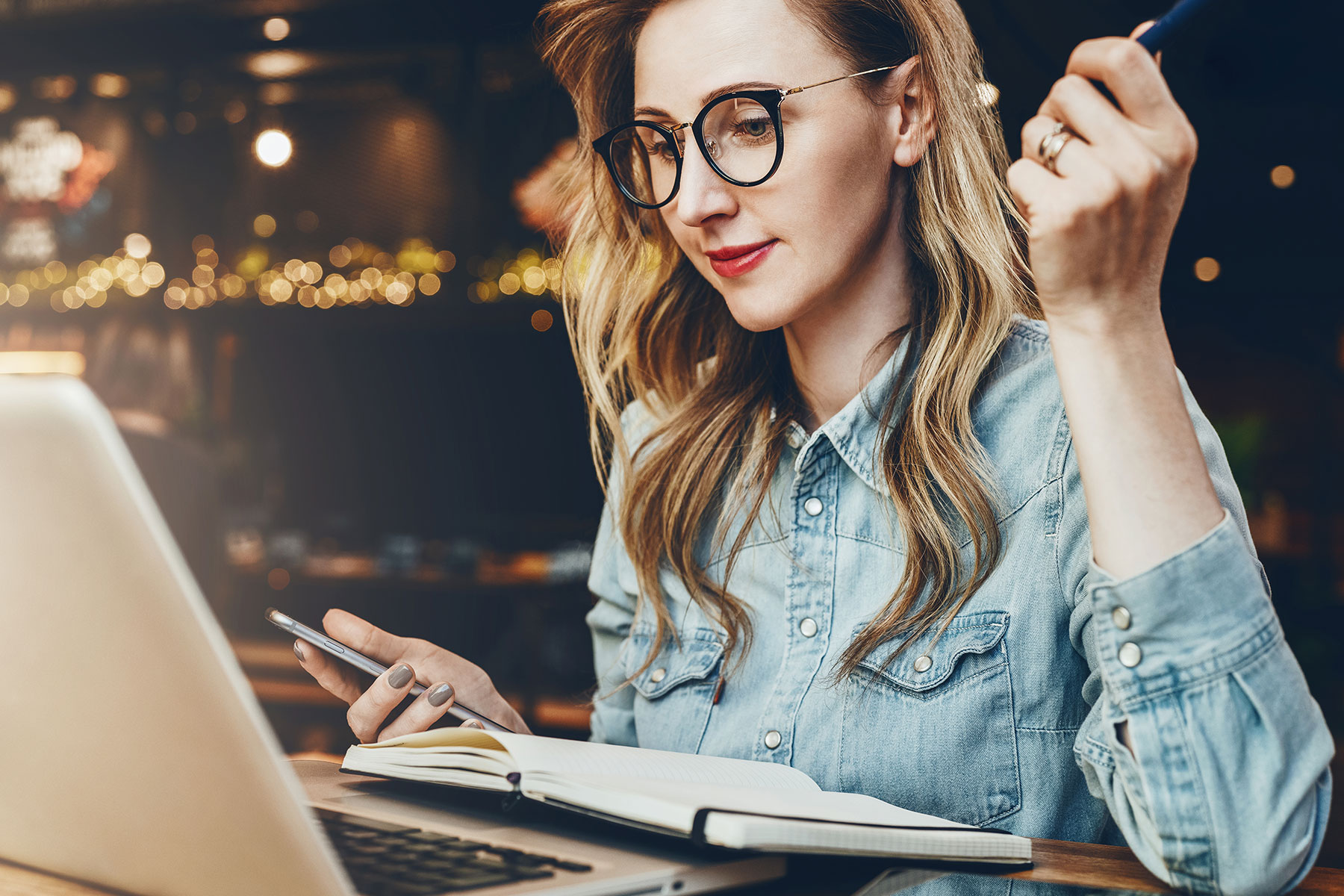 Beautiful young woman wearing think rimmed black glasses, holding a pen and a phone, while looking at a computer