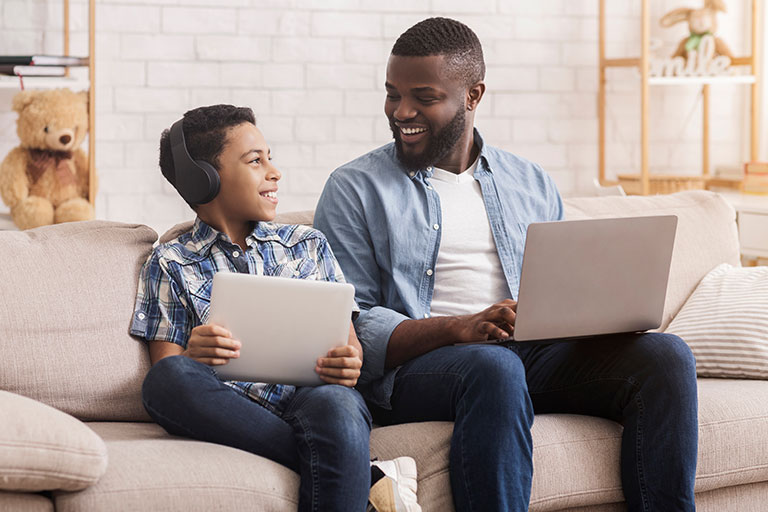 Dad and son sitting on couch with laptop and tablet smiling at each other