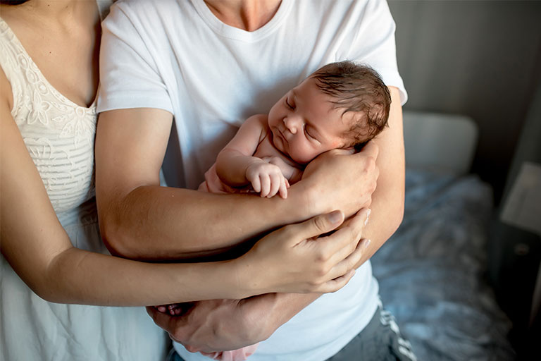 New parents holding baby with dark brown hair in front of window