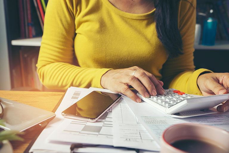 Close up of woman in yellow long sleeve top using calculator with phone and papers in front of her