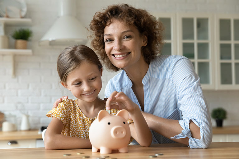Woman with short curly hair and young girl putting coins into piggy bank with sitting at the kitchen counter