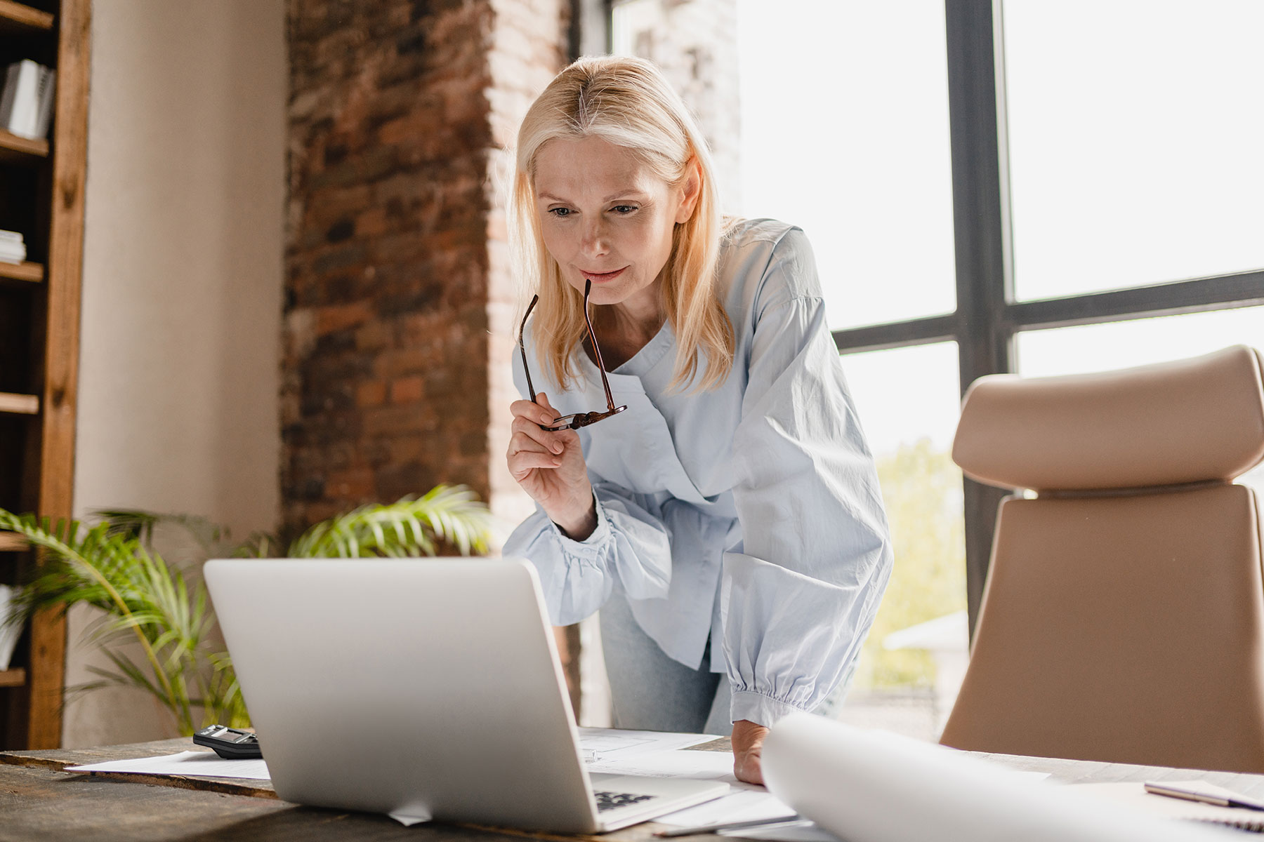 Old causasion woman leaning over desk with earpiece of glasses in her mouth looking at computer