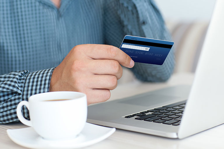 Man in blue plaid shirt holding credit card in front of computer with a cup of coffee 