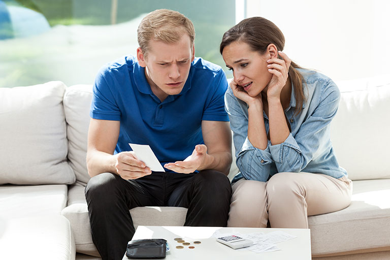 Young couple sitting on couch looking stressed with a wallet, calculator, a change laying on a table in front of them. 