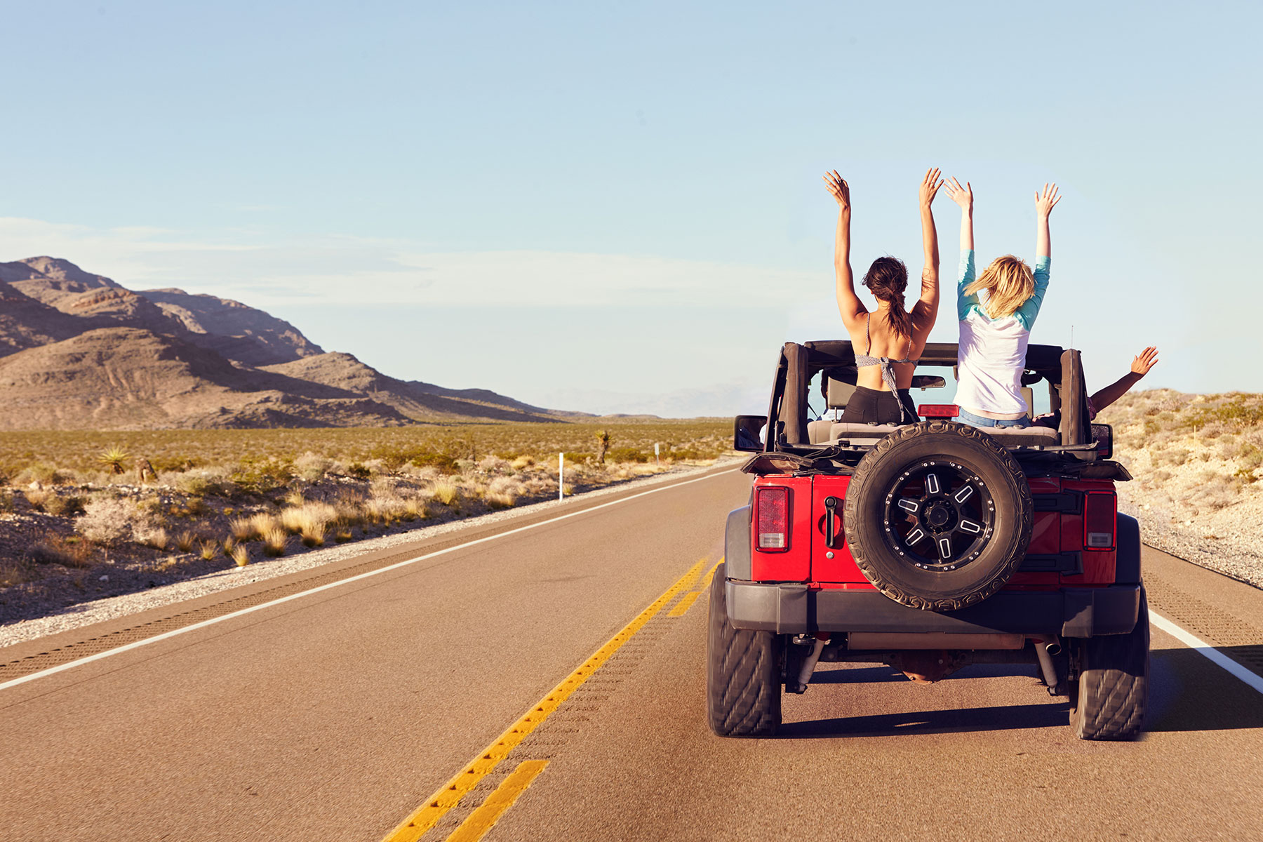 Women sitting on the back of a jeep waving their hands in the air as they drive along a road near mountains