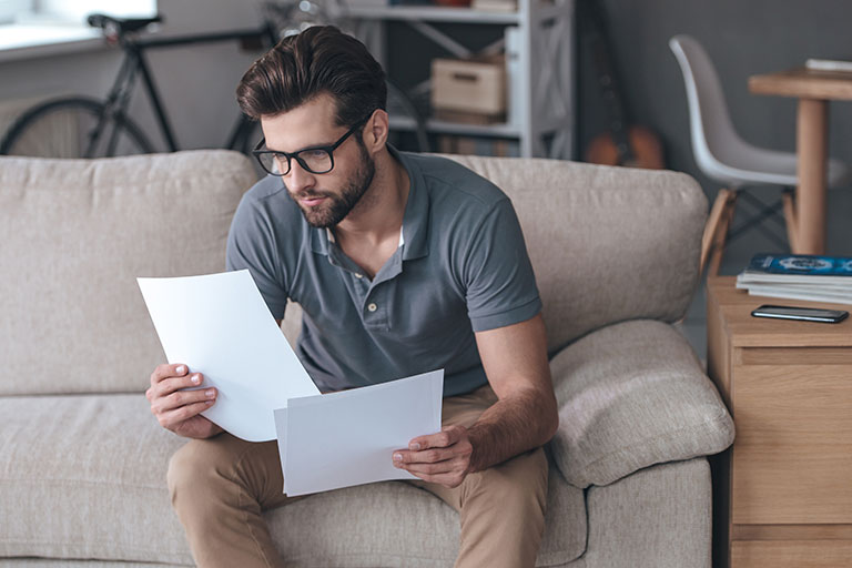 Man wearing glasses and looking at papers, sitting on couch in living room of a home with bicycle and guitar in background