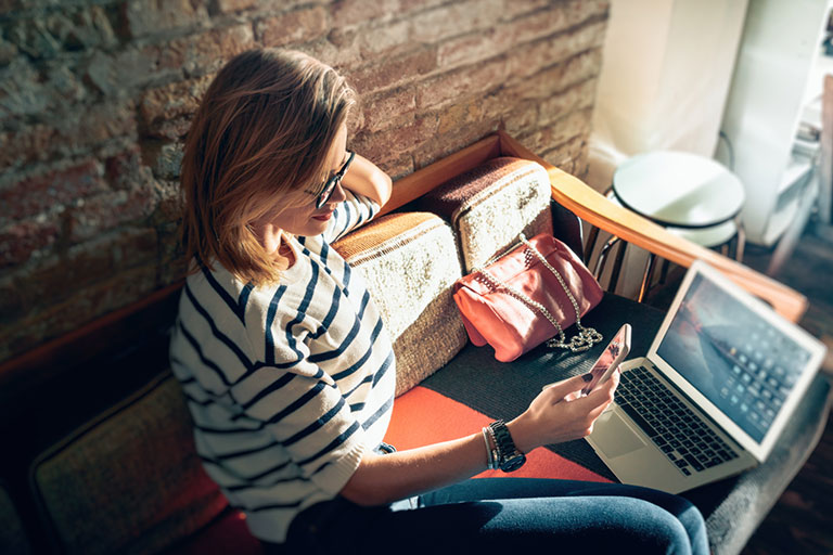 Young woman wearing striped shirt chatting on cell phone while sitting front open computer in vintage coffee shop.