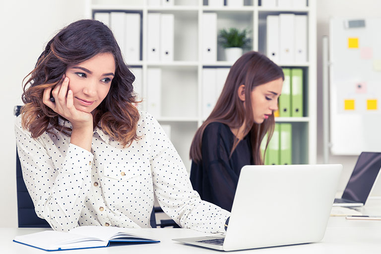 Woman in polka dot blouse resting chin on hand, looking at a laptop computer, another woman looking at laptop in background