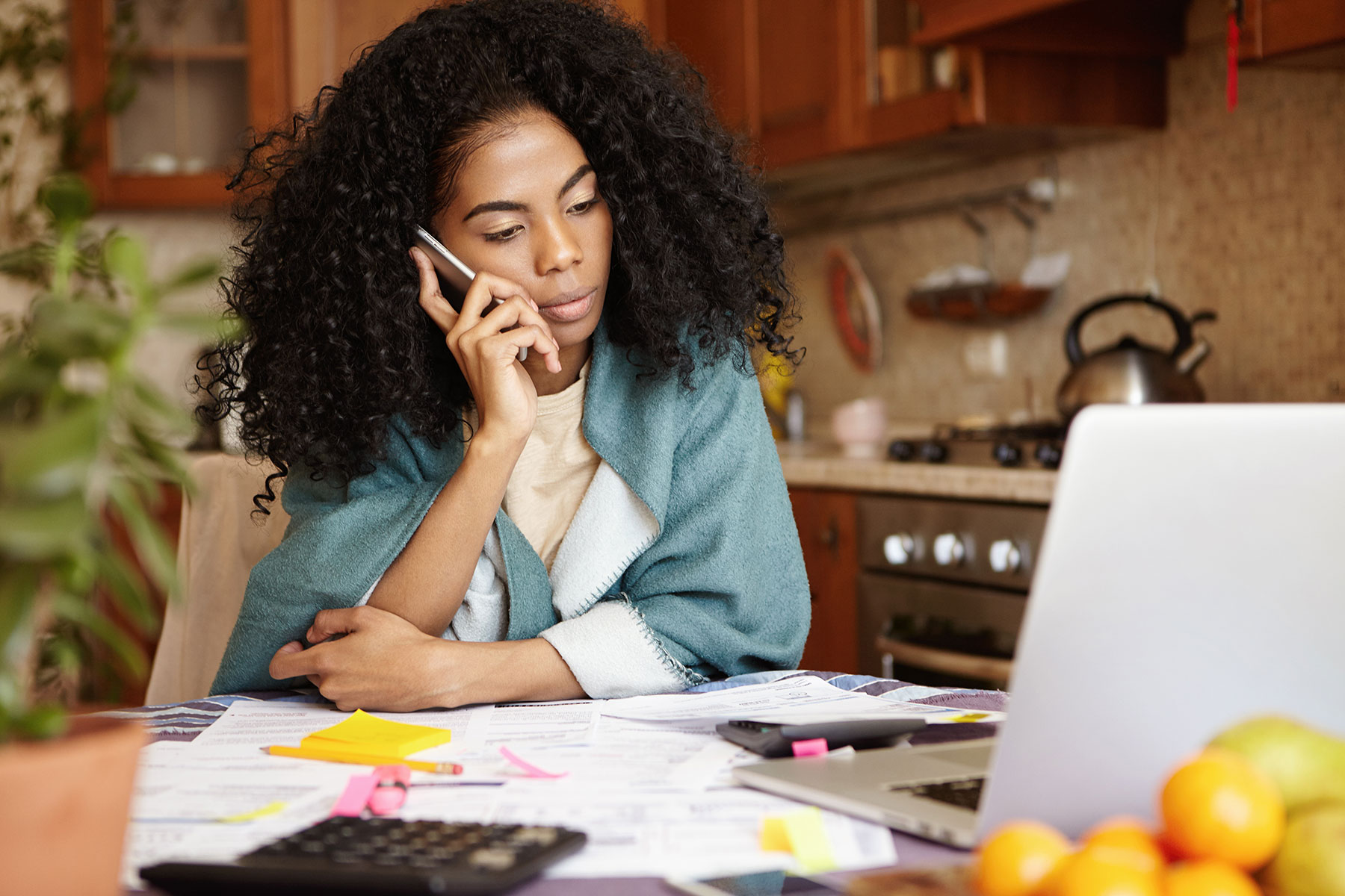 Sad woman sitting in kitchen in front of laptop, talking on mobile phone surrounded by paperwork