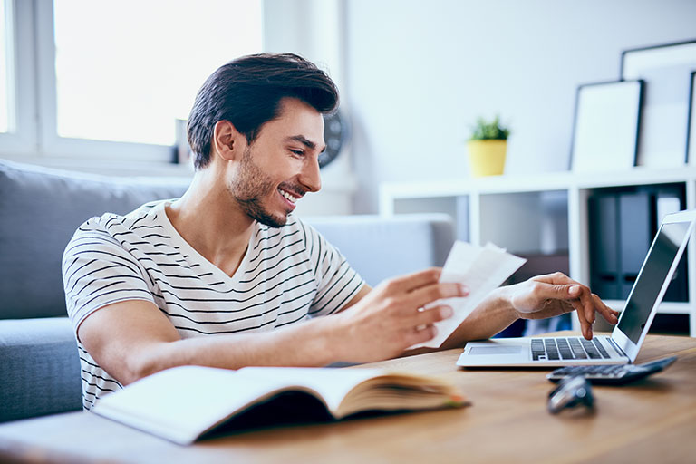 Young man smiling while seated on floor in front of coffee table working on laptop while holding paper