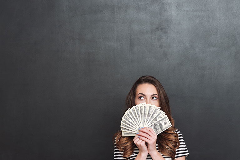 Young brunette woman in front of black background holding fanned out cash and wearing a black and white striped shirt