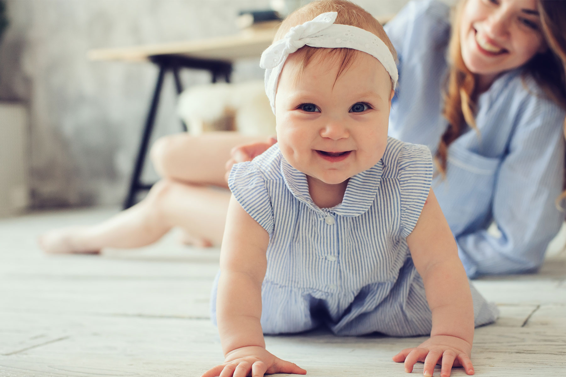 Young baby girl crawing in front of mom wearing a white headband and blue dress
