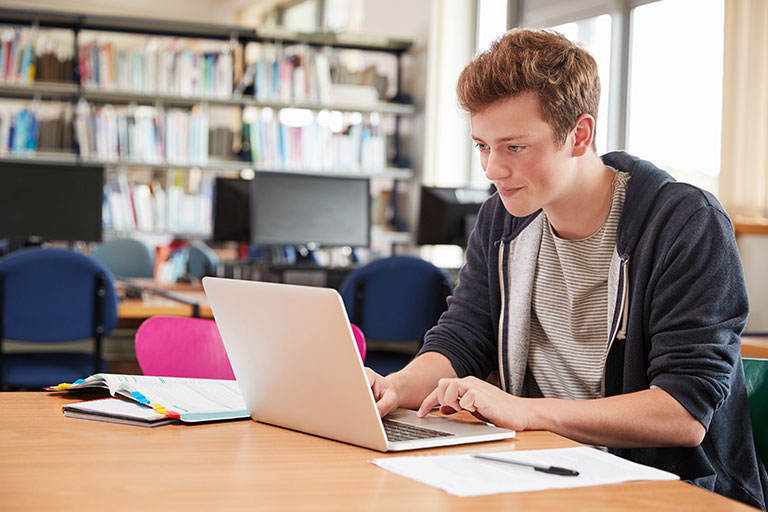 Boyish young man sitting at a desk in a classroom working on his computer.