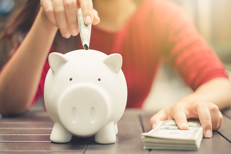 Woman with a stack of cash putting a one dollar bill into a white piggy bank