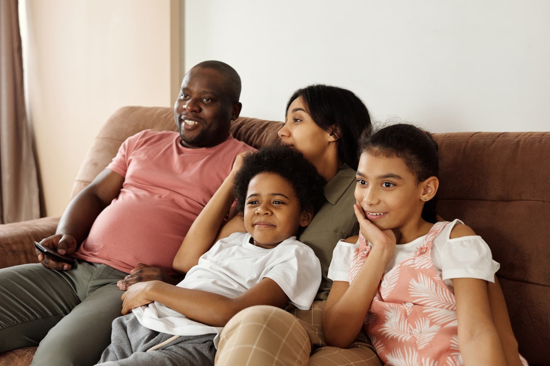 Family lounging on a brown couch together watching tv while the dad holds the remote